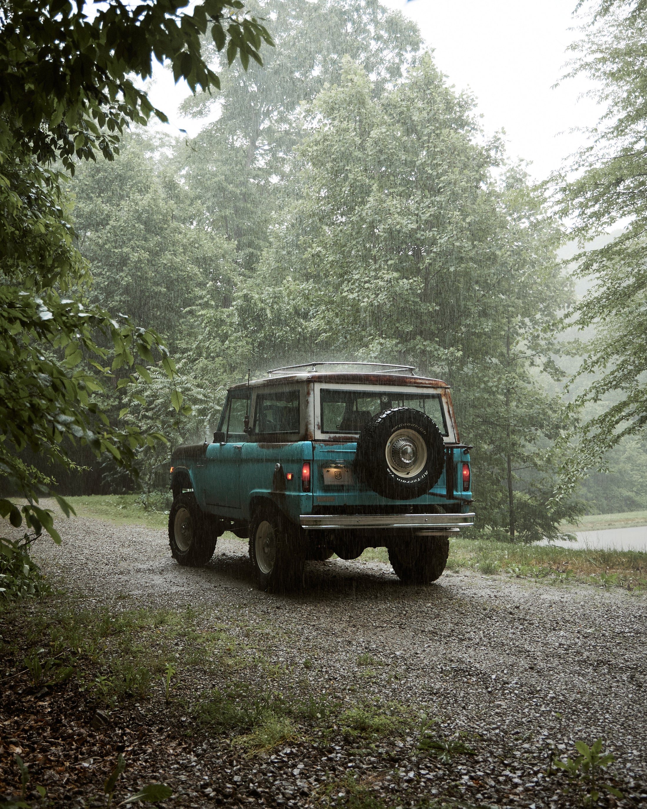 Classic Ford Bronco parked on gravel road in rainy wooded setting