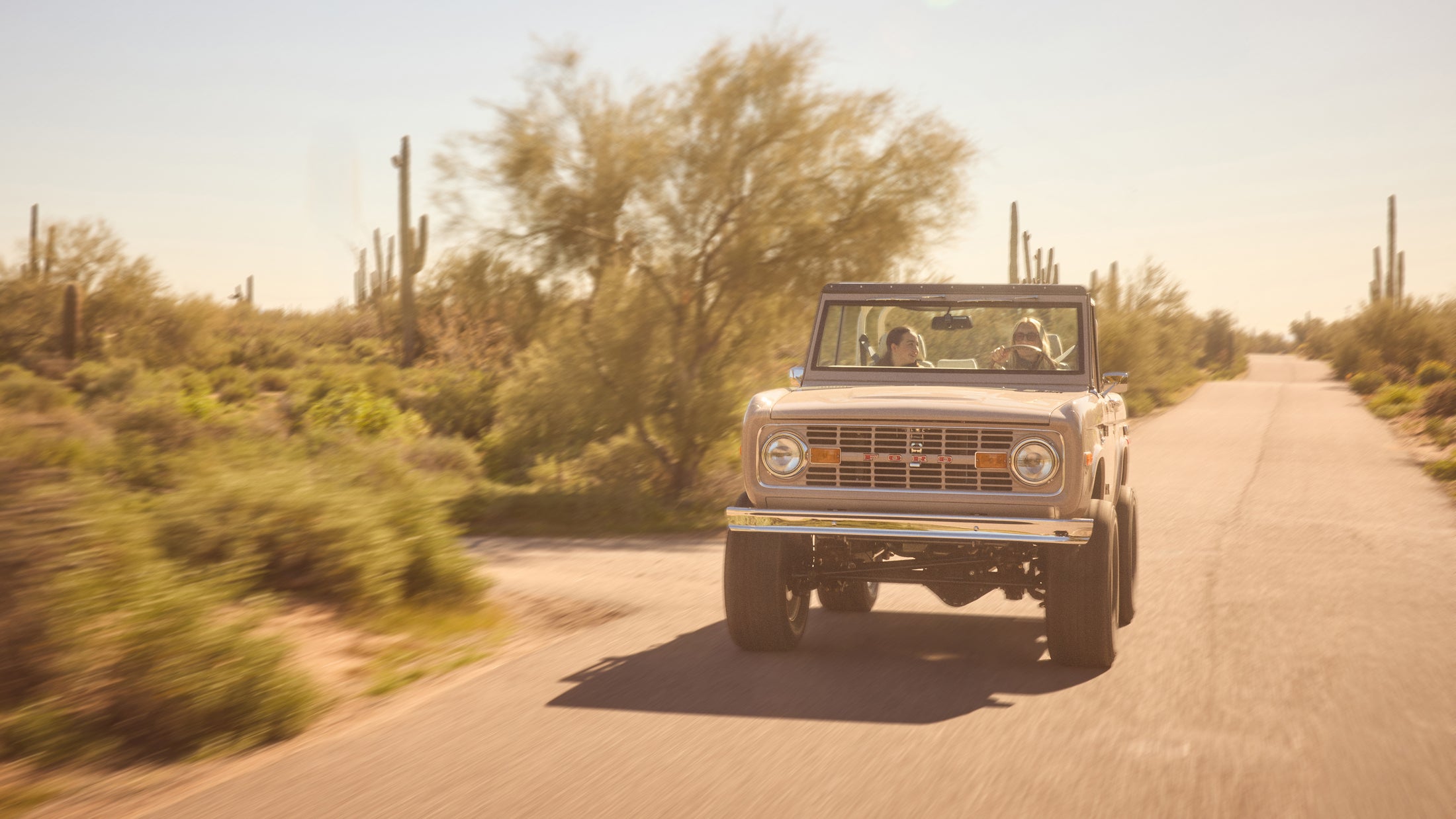 Classic Ford Bronco driving on a desert road with two people inside during golden hour