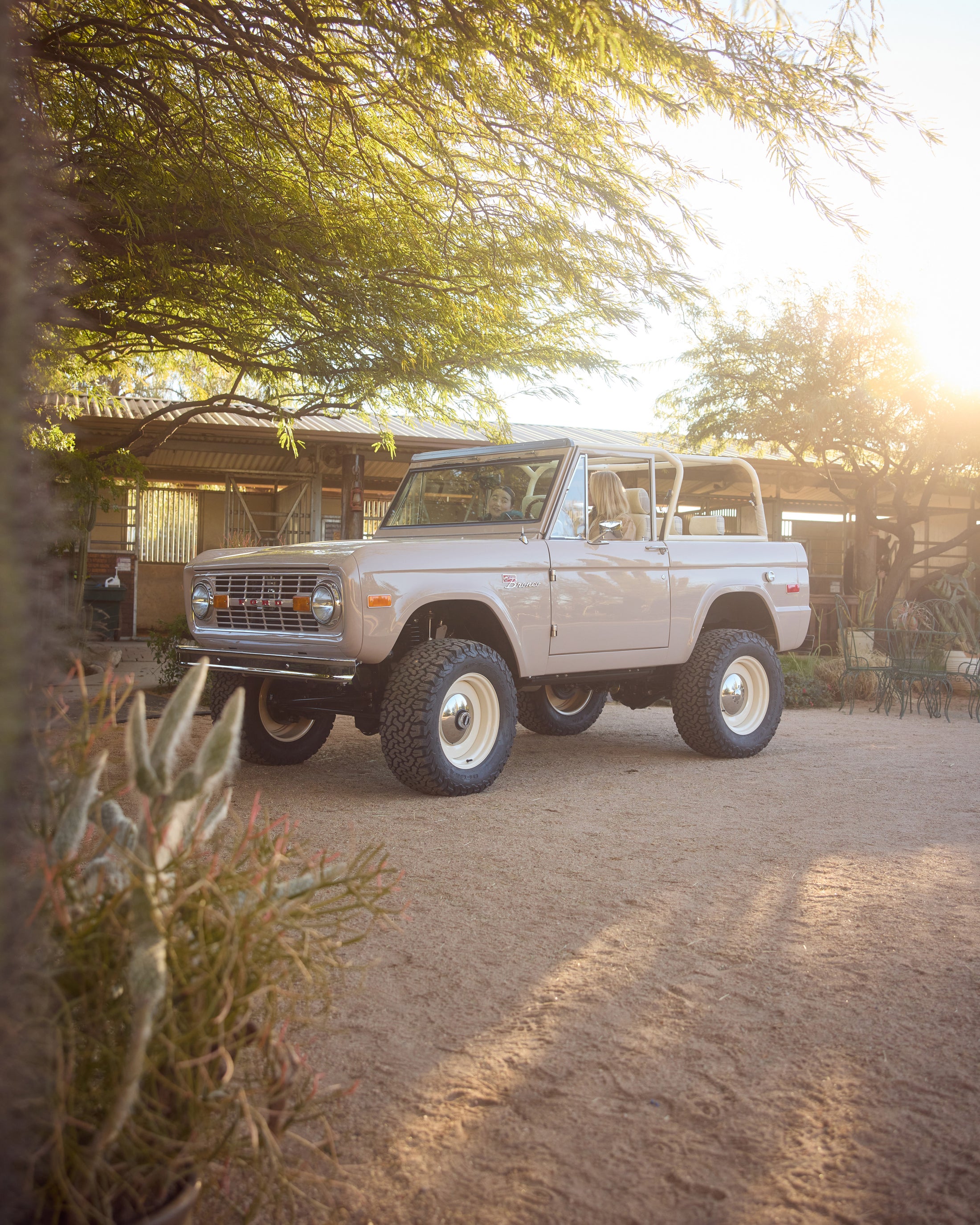 Classic Ford Bronco parked on a sunlit dirt driveway surrounded by desert vegetation