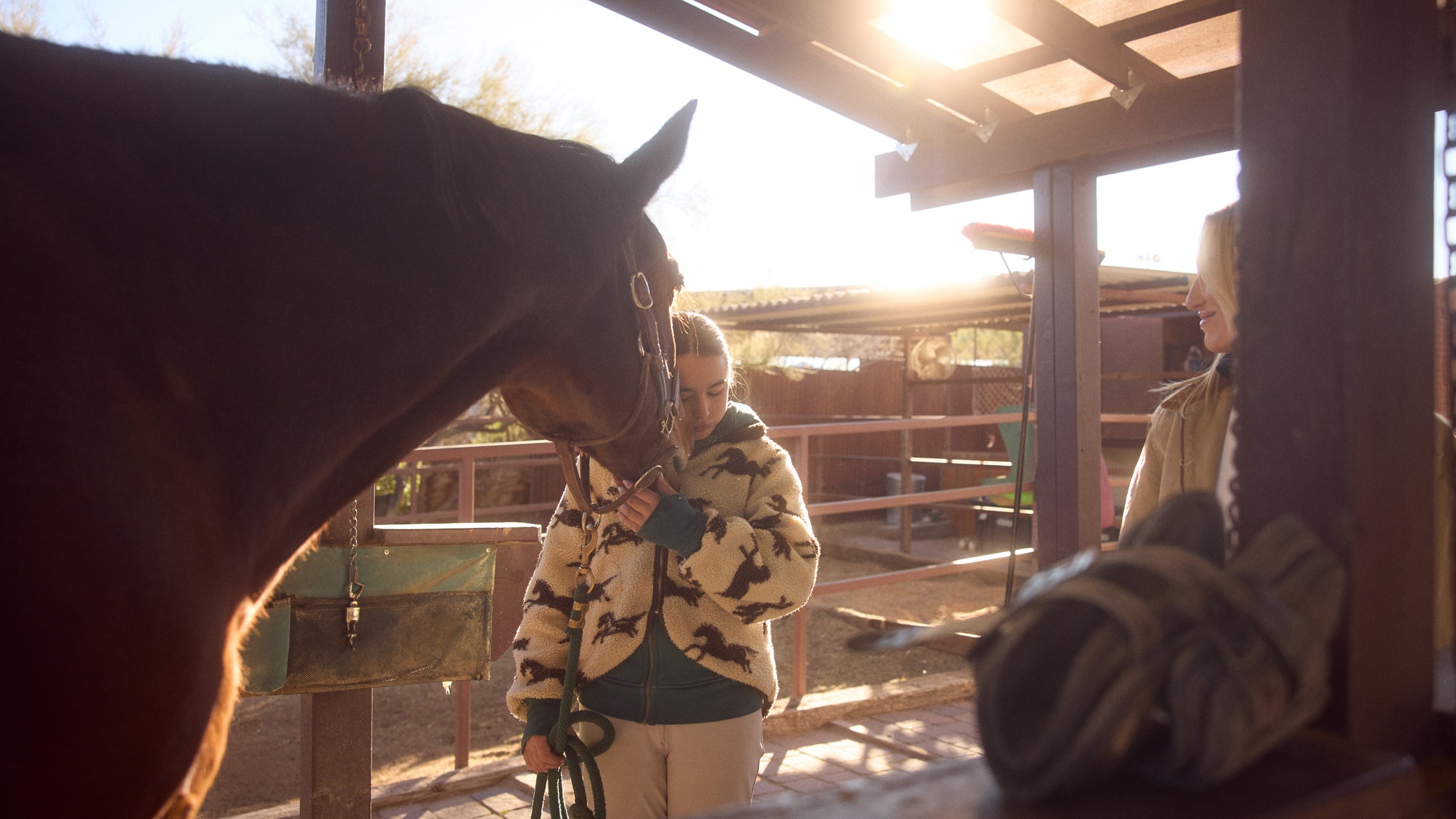 Woman in patterned jacket standing close to a horse under a roofed stable