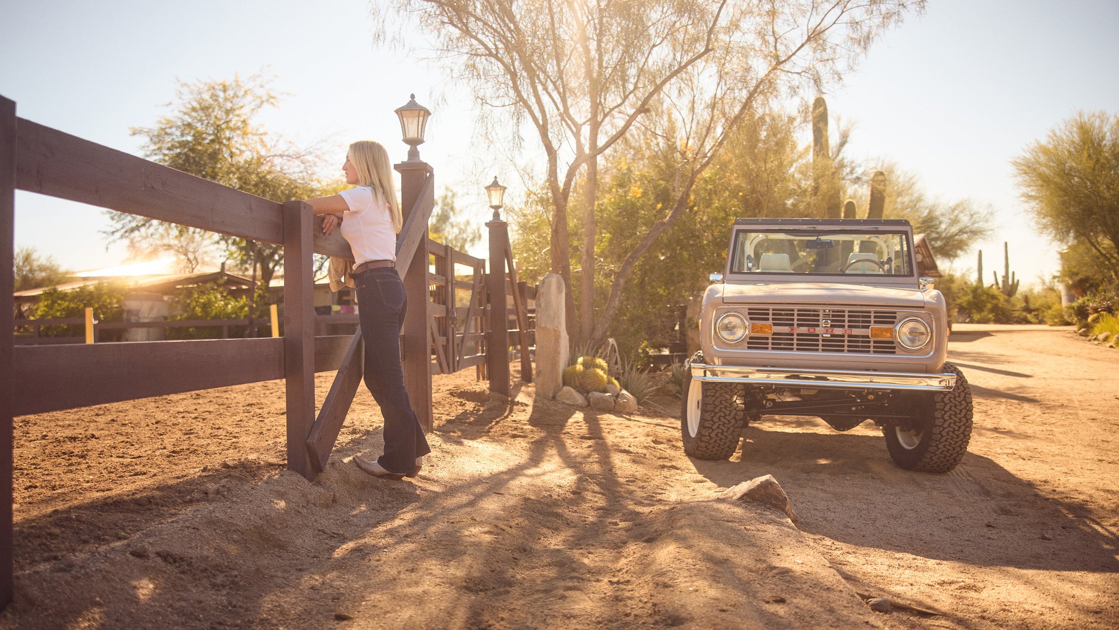 Classic Ford Bronco parked on a dirt road near a wooden fence with a woman leaning on the fence