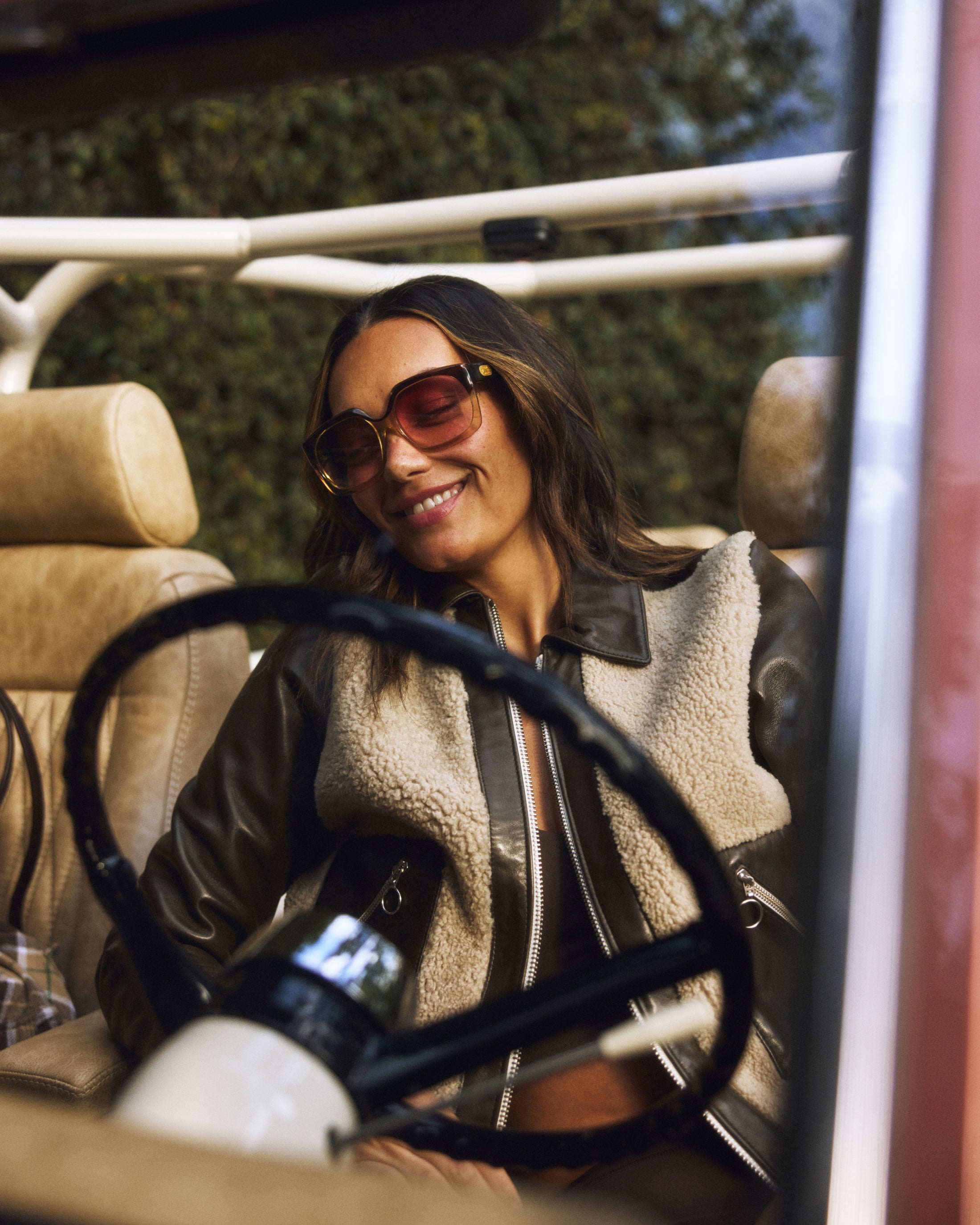 Woman wearing sunglasses and a shearling leather jacket sits relaxed inside a vehicle with tan leather seats and a visible