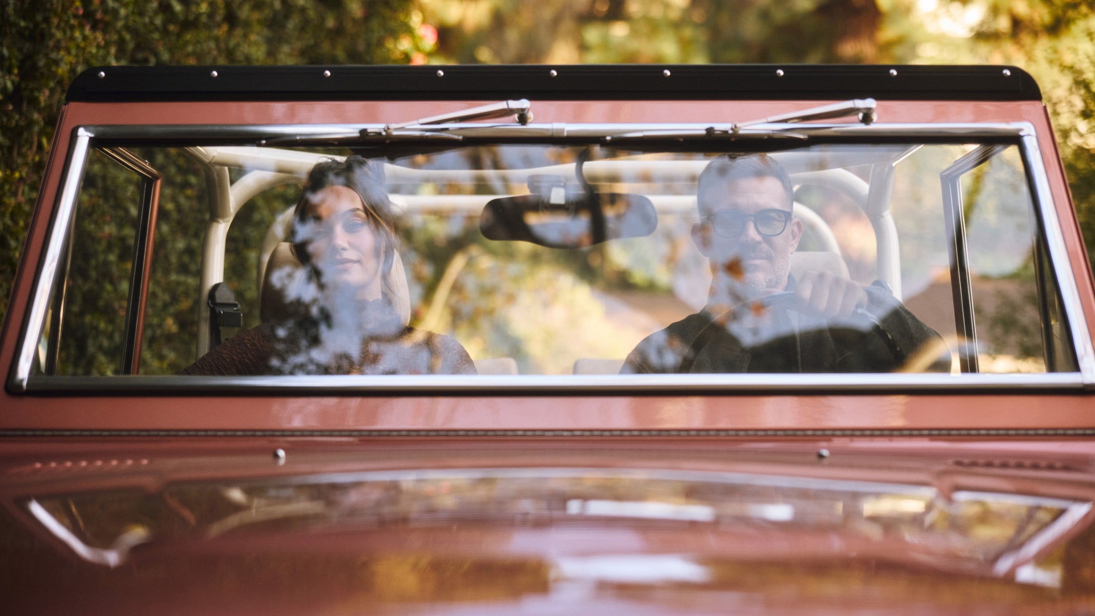Two people seated inside a rust-colored Classic Ford Bronco with autumn foliage reflected on the windshield.