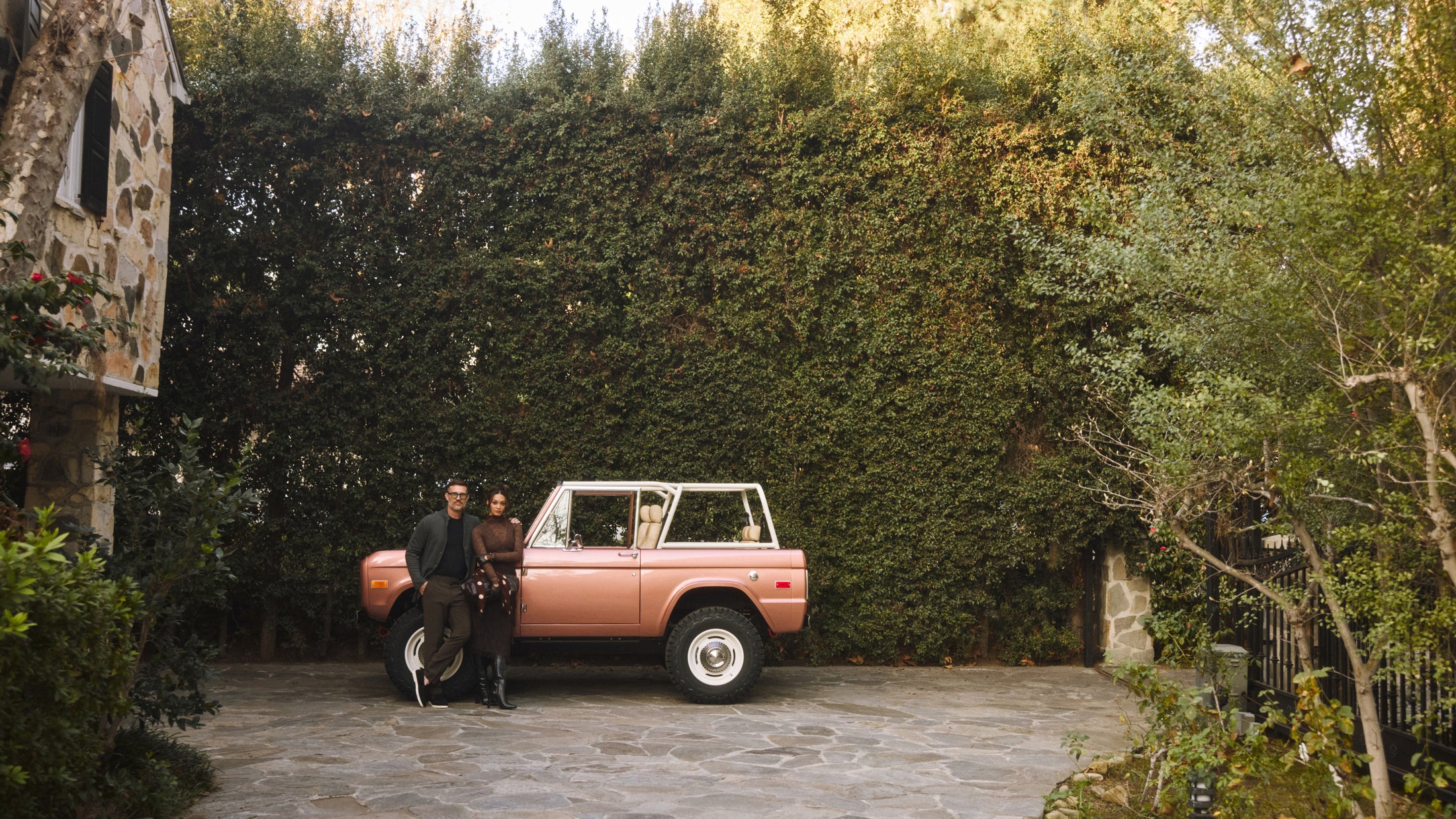 Classic Ford Bronco parked on a stone driveway with two people standing beside it and a tall hedge in the background.