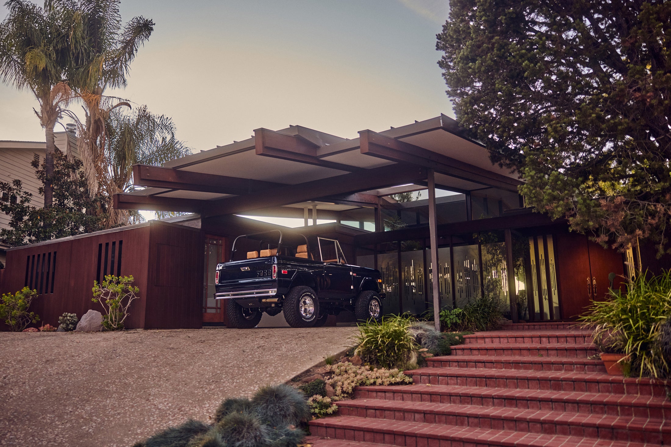 Black Classic Ford Bronco parked in front of a mid-century modern house at dusk.