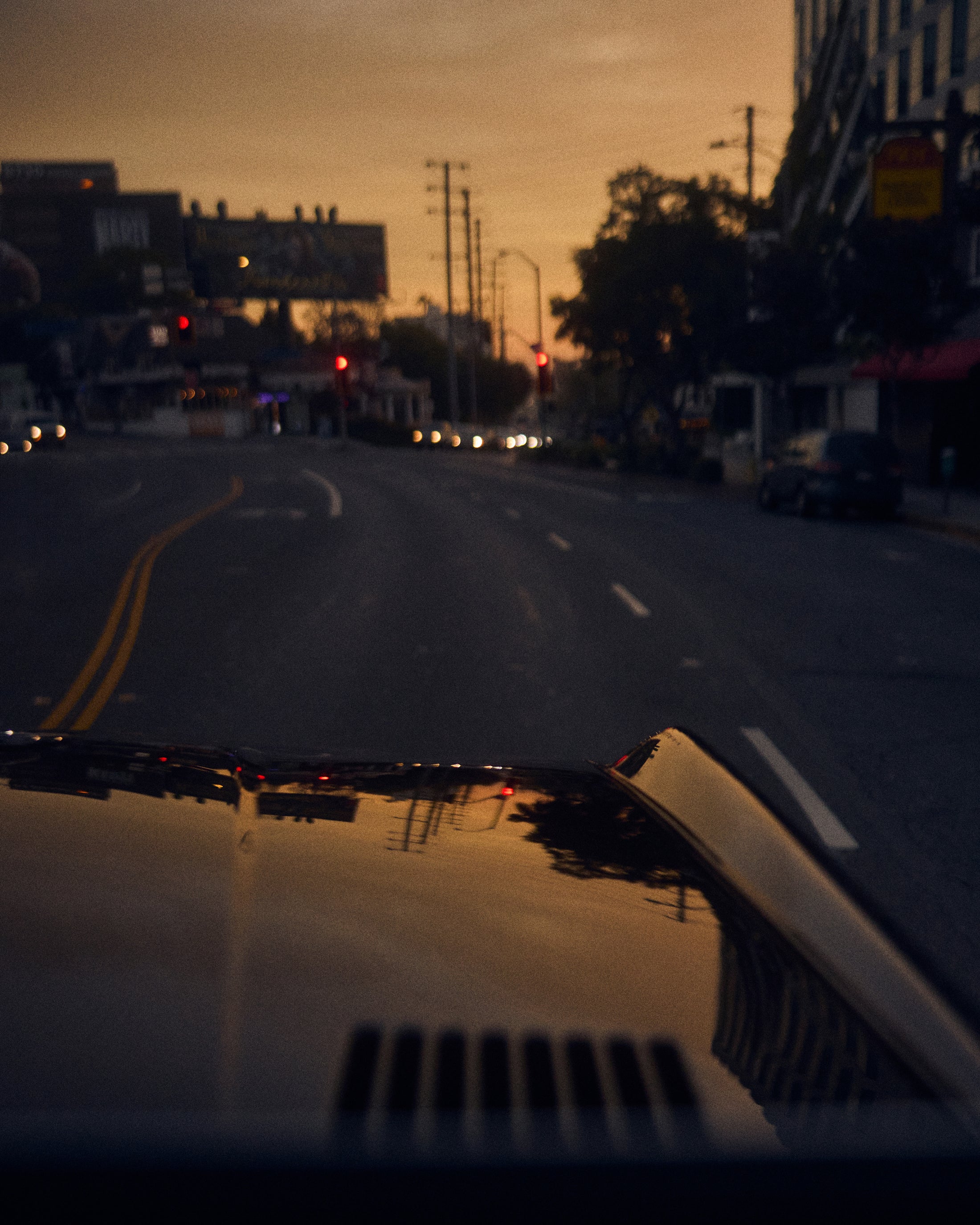 View over the hood of a Classic Ford Bronco driving on a street at dusk with traffic lights ahead.