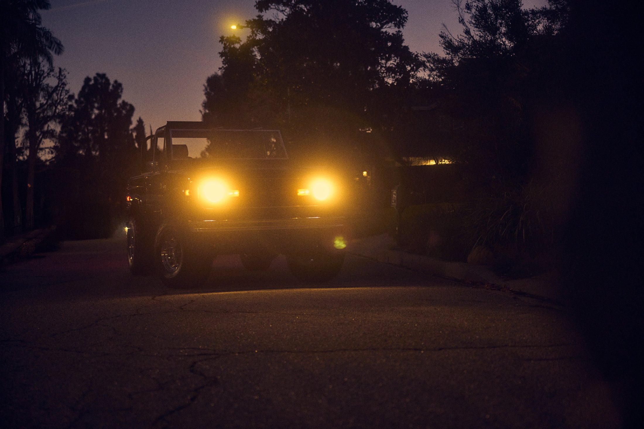 Classic Ford Bronco parked on a twilight residential street with headlights on.