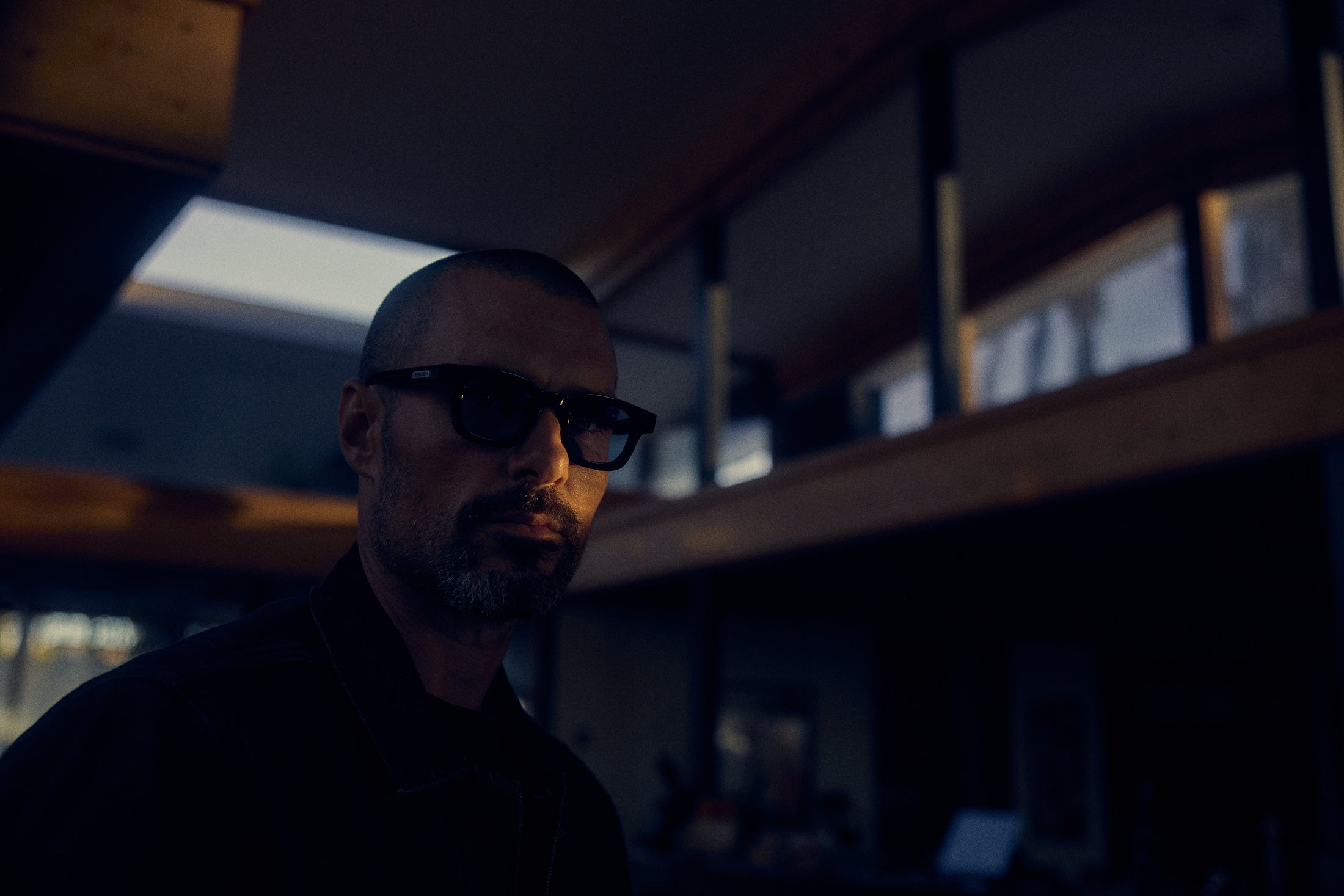 Man wearing sunglasses in a dimly lit room with modern interior design and windows overhead.