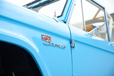 Side front angle view showing door and mirror of light blue Classic Ford Bronco with beige roll cage and seats