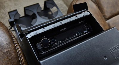 Black console housing a Kenwood stereo unit with cup holders and brown leather seats nearby in Classic Ford Bronco
