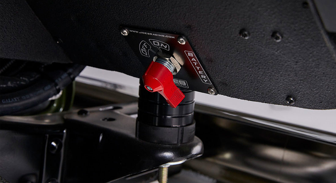 Red battery disconnect switch mounted on black surface beneath olive green Classic Ford Bronco body