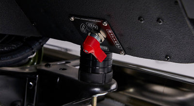 Red battery disconnect switch mounted on black surface beneath olive green Classic Ford Bronco body