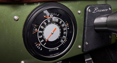 Classic Ford Bronco speedometer with black dial, white numbers, and orange needle on green dashboard