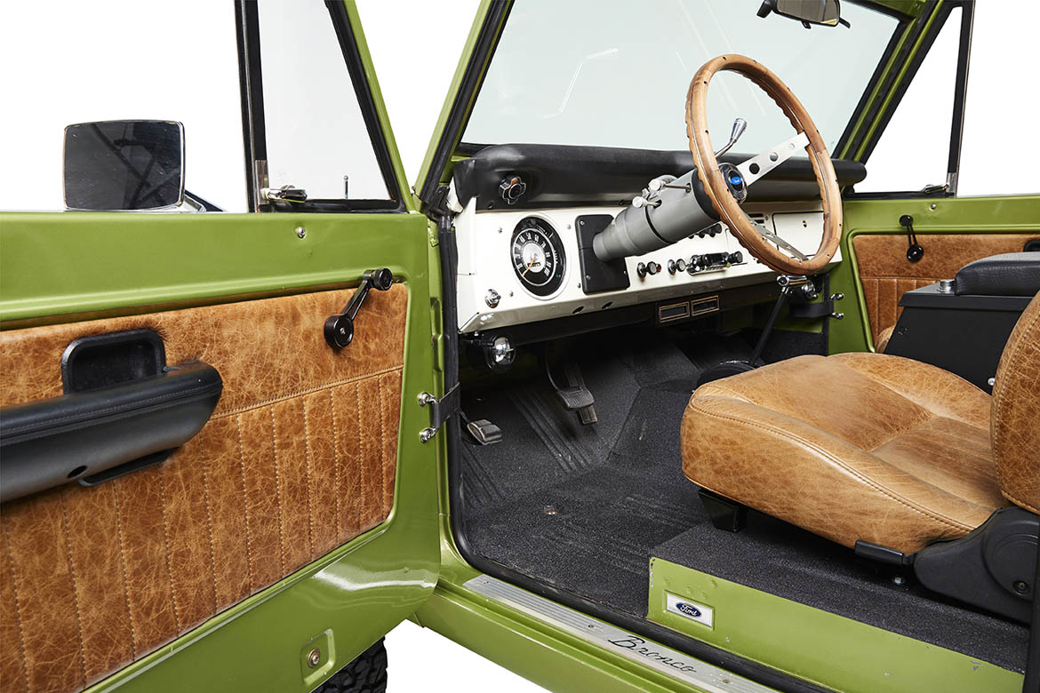 Open driver side door of Classic Ford Bronco showing steering wheel, dashboard, and brown leather seats.