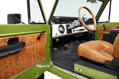 Open driver side door of Classic Ford Bronco showing steering wheel, dashboard, and brown leather seats.