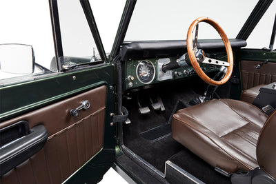 Classic Ford Bronco driver-side interior showing brown seats, door panel, silver pedals, and wooden steering wheel.