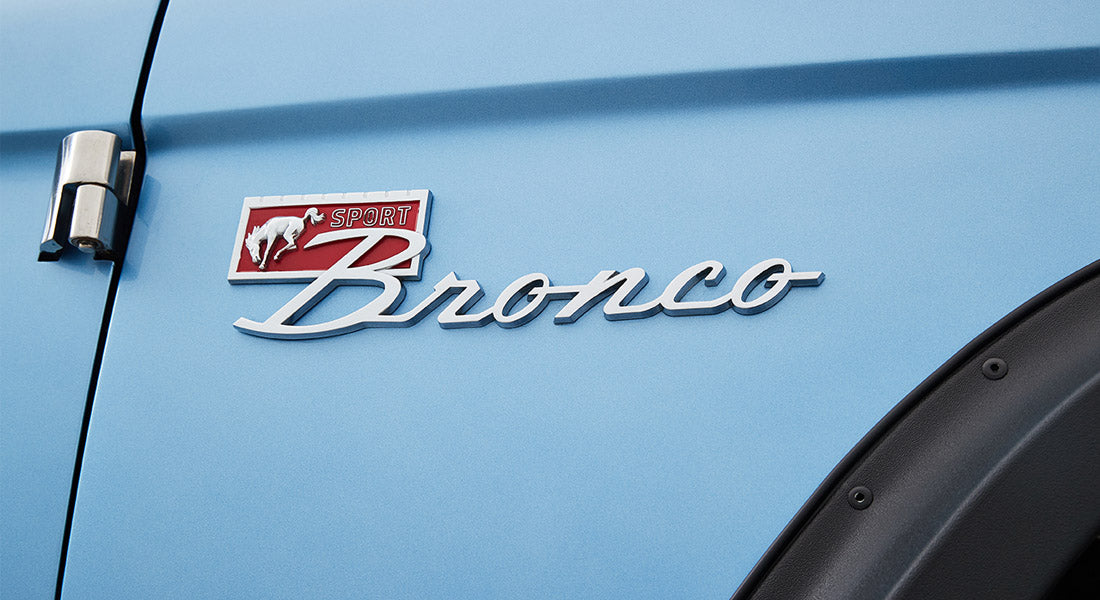 Classic Ford Bronco emblem badge on light blue exterior door panel with black fender flare.