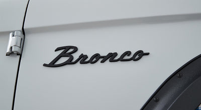 Matte black Classic Ford Bronco emblem on white vehicle door near hinge
