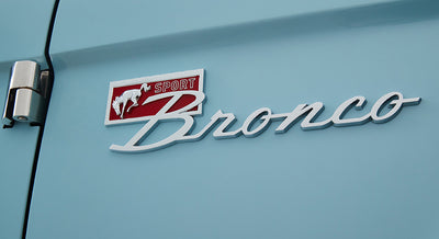 Classic Ford Bronco chrome emblem with red and white background on light blue vehicle body.