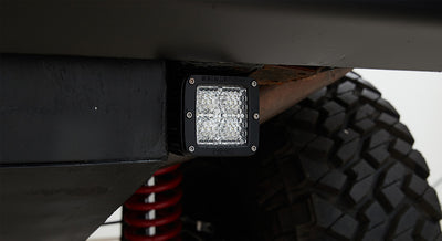 Square LED light mounted on black frame near large off-road tire and red coil spring suspension