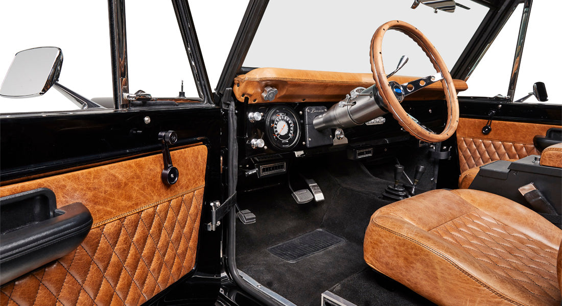 Classic Ford Bronco interior showing brown leather steering wheel, dashboard, pedals, and quilted brown panels