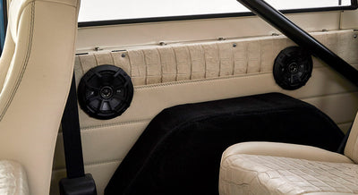 Two black round speakers mounted on tan leather interior panel behind seat with roll bar