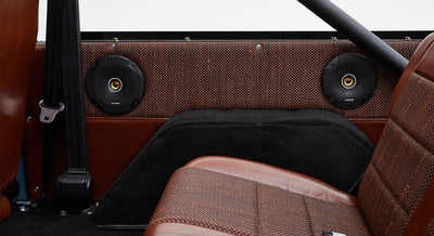 Speakers mounted on the interior side panel behind a brown leather car seat with woven detailing.