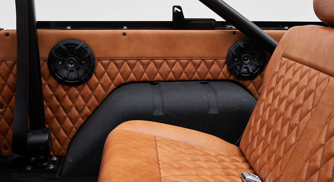 Two black speakers mounted in quilted brown interior door panel behind tan seat inside Classic Ford Bronco