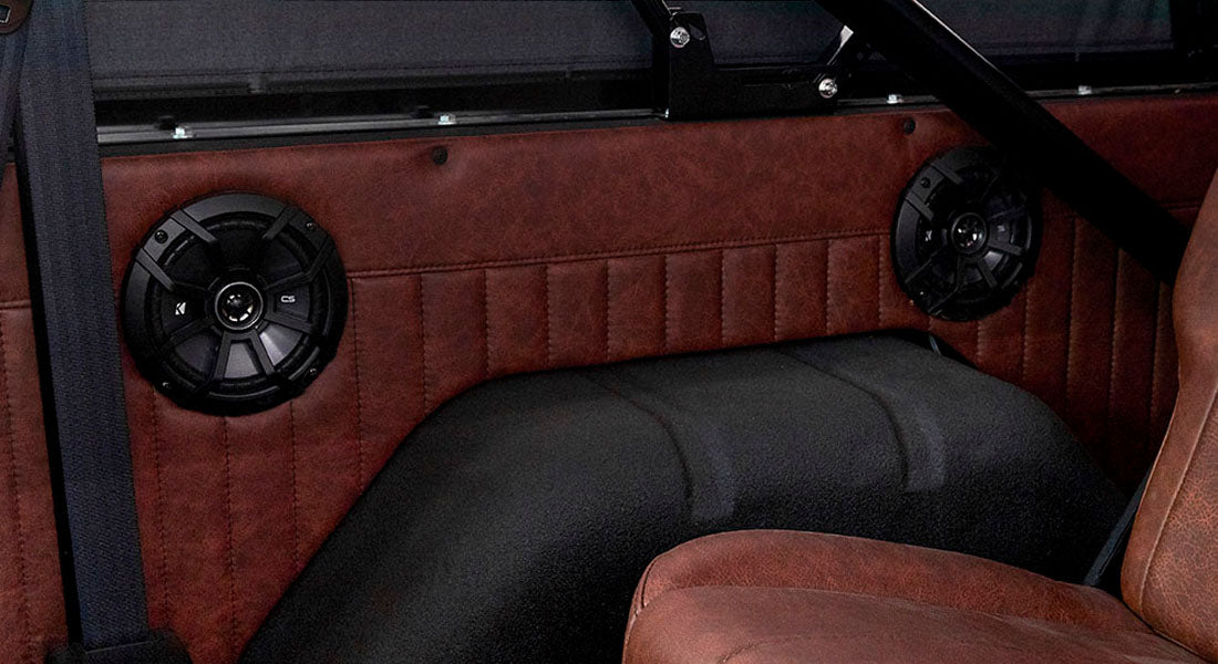 Two black speakers mounted on reddish-brown leather interior panel behind vehicle seat