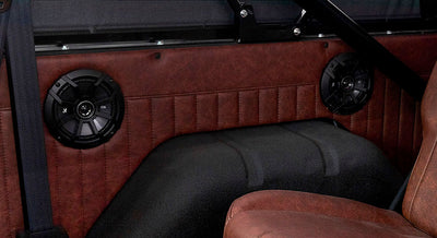 Two black speakers mounted on reddish-brown leather interior panel behind vehicle seat