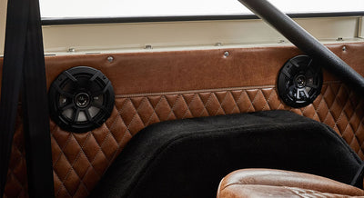 Two black round speakers mounted on quilted brown leather interior panel with part of a steering wheel and seat visible.