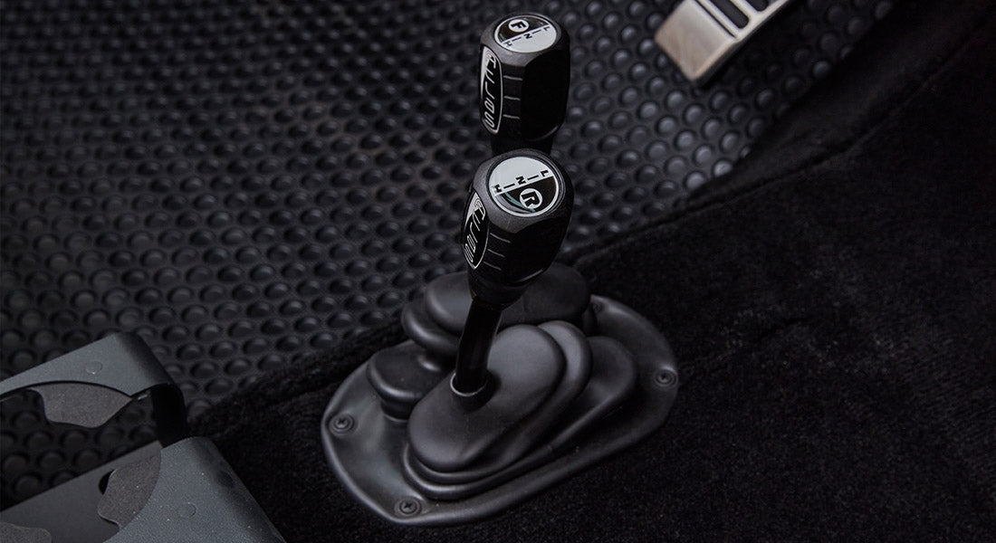 Two black transfer case shifters with white markings on a black rubber floor mat