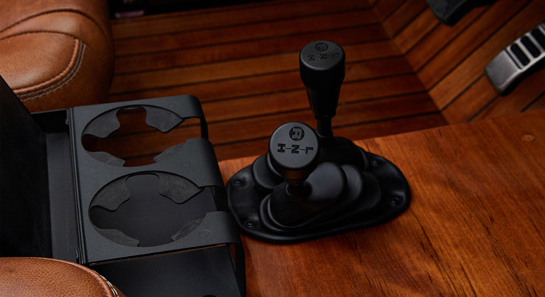 Two black gear shift levers on wooden floor next to black cup holder in Classic Ford Bronco